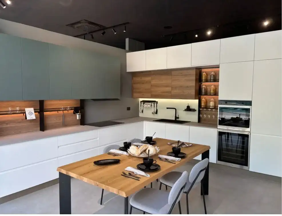 Cucina da esposizione bianca con ante in rovere, pensili verde salvia e tavolo in legno, in showroom